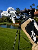 A beige and black stand bag filled with silver and white golf clubs, set on a golf course with a pond, green grass, and palm trees in the background.